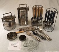 Photo showing two stainless steel tiffin boxes, black-handled cutlery set on stand, cream-handled knives, vintage tins, miscellaneous cutlery and lemon juicer.