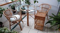 Overall view of round bamboo and glass table with two bamboo chairs around it in a tiled room with plants.