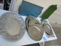 Photo showing the complete lot of 9 large catering trays arranged on a white table. The large Wilton pewter look tray with fruit border is visible along with several aluminum trays and a green vintage plastic tray with Roman details.