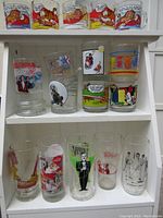 Three-tier shelf displaying the full lot of 17 glasses: top row Garfield tumblers; middle row Star Trek III stein, Olympic mug, Norman Rockwell cowboy, Viva Variety plastic, Garfield and Mickey; bottom row Bicentennial, Coca-Cola Santa and additional Rockwell designs.