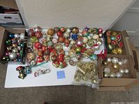 Full overview of about seventy vintage decoupage and goldtone glass Christmas ornaments arranged on table and in boxes, showing variety of shapes, colors, and styles.