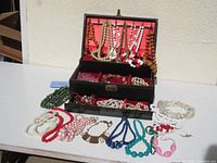 Open black jewelry box with red interior showing multiple beaded necklaces hanging from lid and compartments filled with various bead necklaces, some spilling onto table surface.