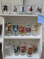 Photo showing three shelves with 15 glasses total, including Norman Rockwell, Bugs Bunny, Pepsi promotional and Christmas themed glasses