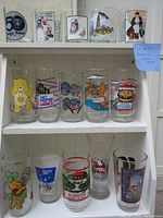 Shelf display of 15 assorted vintage promotional glasses with colorful cartoon and brand graphics