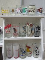 Front view of 16 promo glasses arranged on a white shelf showing various cartoon and souvenir glass designs including Disney, Warner Brothers, Jelly Jar, and others