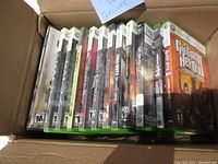 Photo shows a box containing 19 Xbox and Xbox 360 game cases arranged side by side showing their spines for title identification.
