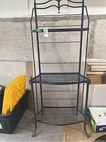 Front view of black metal baker's rack showing overall structure, three slatted shelves, and decorative upper back panel.