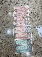 Nine Canadian banknotes laid out on a surface, showing two one-dollar bills at the bottom and seven two-dollar bills above them, some with color tones of green and pink/red.
