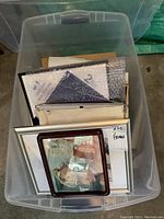 Plastic bin containing assorted picture frames stacked vertically, various sizes and types including brown, black, silver, and wooden frames with glass and backing