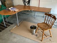 Photo showing L-shaped wooden Ikea sectional desk with silver metal legs in a garage setting, with wooden chair and metal basket in foreground.