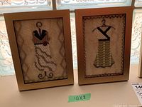 Two framed textile art pieces showing stylized dresses on hangers with beadwork and embroidery, labeled 10x8 inches.