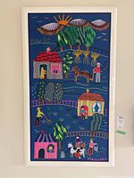 Full view of framed textile art showing colorful embroidered rural countryside scene with houses, people, animals, trees, fences and mountains in background.
