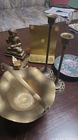 Brass figurine sitting on a wooden surface along with candlesticks, bowl, basket, and book ends scattered around it.