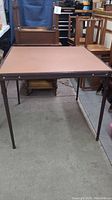 Side view of a square card table showing metal legs and brown vinyl top with some visible marks and wear.