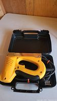 Yellow Arrow electric staple applicator stapler with power cord inside black plastic carrying case, shown from top and side views.