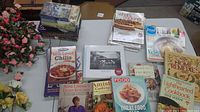 Full view of books, cookbooks, and magazines laid out on table alongside faux floral arrangements with pink and yellow flowers.