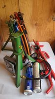 Photo showing green metal car stands, jumper cables, engine compression gauge, and two cans of grease with applicator tool on a white table in wooden room.