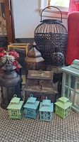 Overview of twelve assorted decorative lanterns including a large black birdcage-style lantern, a house-shaped lantern with Asian characters, several medium-sized metal lanterns with patterns, and four small pastel green and blue lanterns in front.