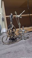 Four metal musician sculptures including cyclist, flute player, violin player, and seated figure, displayed together on floor in front of wooden shelves.