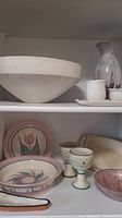 Shelved display of pottery items including large beige bowl, tall vase, goblets, decorative bowls with floral designs, cream and sugar dish and tray.