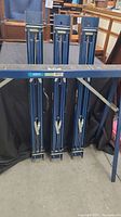 Four folded Mastercraft blue metal saw horses leaning against a wall showing worn and used condition.