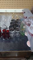 Full lot of assorted stemware shown on table with power outlet backdrop. Various glasses including pink champagne coupes in boxes, clear wine glasses, dark red brandy glasses, and other small stemware.