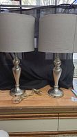 Pair of table lamps shown side by side on a wooden surface with black background, showing metal bases and grey fabric drum shades, electrical cords attached.
