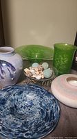 Five glass items on floor including green vase, green bowl, blue bowl, purple brushstroke vase, pink-white vase and metal stand with decorative orbs.