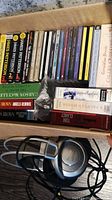 Box showing multiple audiobook CDs and boxed audiobooks by various authors including Dan Brown, James Patterson, Stuart McLean, and Tom Clancy along with Panasonic RP-HT770 headphones lying in the box