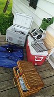 Image showing a gray Coleman thermoelectric cooler opened with cables inside, red and white Coleman Polylite54 cooler closed, black and white portable music player, folded green/black chairs and blue inflatable mattress with pump on a porch wooden floor, plus a woven picnic basket with plastic dinnerware.