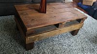 Front angle view of the rustic handcrafted coffee table showing the plank top and sturdy legs on a speckled carpet.