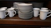 Multiple stacks of white dinner plates and side plates along with white mugs with gold rim displayed on a wooden shelf.