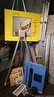 Photo showing aluminum adjustable easel with a yellow Strathmore sketchpad attached and some art supplies on the floor