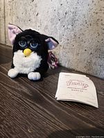 Full view of black and white Furby with yellow beak, blue eyes with glasses, tags attached and instruction manual beside it