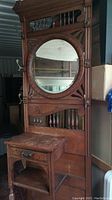 Front full view of wooden hall tree, showing round beveled mirror, small dovetailed drawer, brass hat hooks, umbrella port base