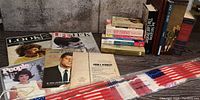 Overview of books and magazines on US political figures with JFK focus, plus folded US flag in packaging on table.