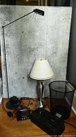 Overview of five items: black adjustable floor lamp, silver table lamp with cream lampshade, magnavox projection alarm clock, black mesh wire wastebasket, APC Battery Backup ES-750 model on wooden floor against wall.