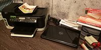 Photo of black HP multi-function printer with front paper tray and some printed pages, Dell Inspiron 1564 laptop closed next to it on desk.