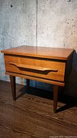 Front and side view of mid-century modern single drawer walnut veneer nightstand showing wood grain, lateral wood pull, and tapered legs.