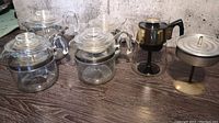 Four glass percolators arranged on wooden floor with visible glass lids, handles, metal bands, and a separate vintage percolator with black handle and metal base