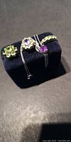 Three sterling silver rings with green and purple semi-precious stones and one sterling silver necklace with purple heart-shaped pendant arranged on black jewelry display.