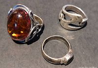 Three sterling silver rings shown on dark surface, one with a large oval amber stone and two plain silver rings with minimal decoration.