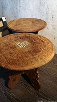 Pair of round, intricately carved wooden tables side-by-side showing tops with inlays and bases.