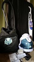 Black H2O branded storage bag, upright white and blue steam mop, and a canister vacuum with a blue translucent water tank. Some accessories and manuals laid out in front.