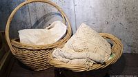 Two natural wicker baskets containing vintage linens with crochet and embroidered floral designs.