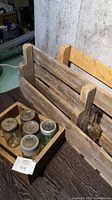 Photo showing two handcrafted wooden shelves with rustic appearance made from wood pallets, one leaning against a wall, the other held upright. Wooden box with Mason jars placed on a dark surface.
