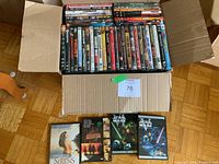 Photo of box open with approximately 80 DVD movies inside showing a variety of titles including classic and modern movies, some Star Wars DVDs visible outside the box on floor.