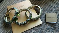 Three Heidi Daus jewelled spring-open bangles displayed on a woven surface with accompanying Heidi Daus card and booklet.