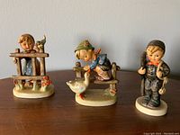 Three Goebel Hummel figurines including a girl behind a fence with a bird, a boy leaning on a fence with a goose, and a standing boy with a bird and accessories on a wooden surface
