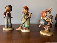Photo of three vintage Goebel Hummel porcelain figurines, depicting children in different activities, placed on a wooden surface.